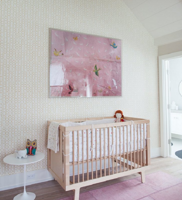 اتاق نوزاد مدرن با کاغذ دیواری طرح شکل هندسی و تخت نوزاد چوبی که با دکوری هایی به رنگ گرم، فرش و تابلو صورتی، دیزاین شده است